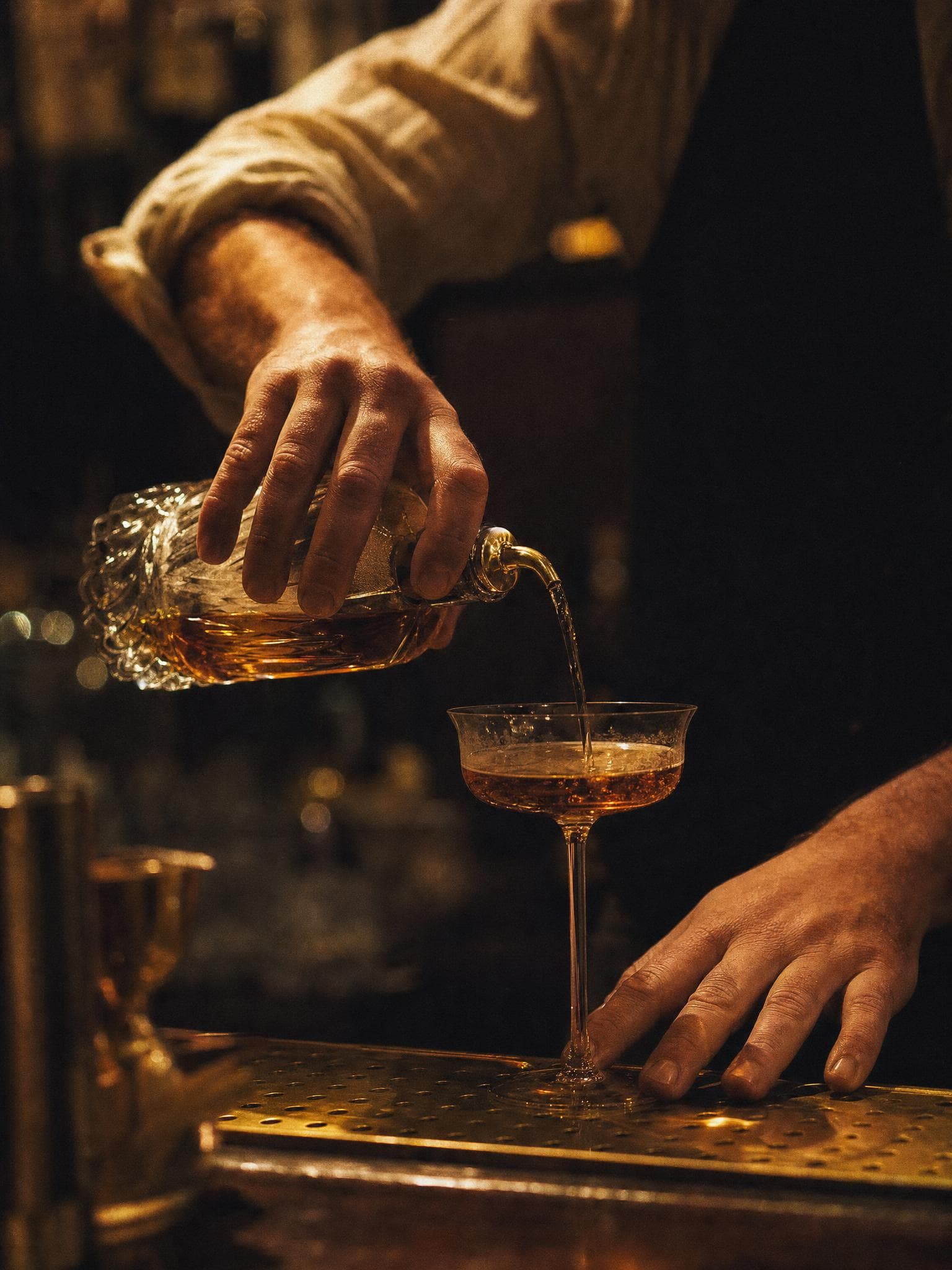 A bartender pours an amber spirit from a cut-crystal decanter into a coupe on a brass bar mat.