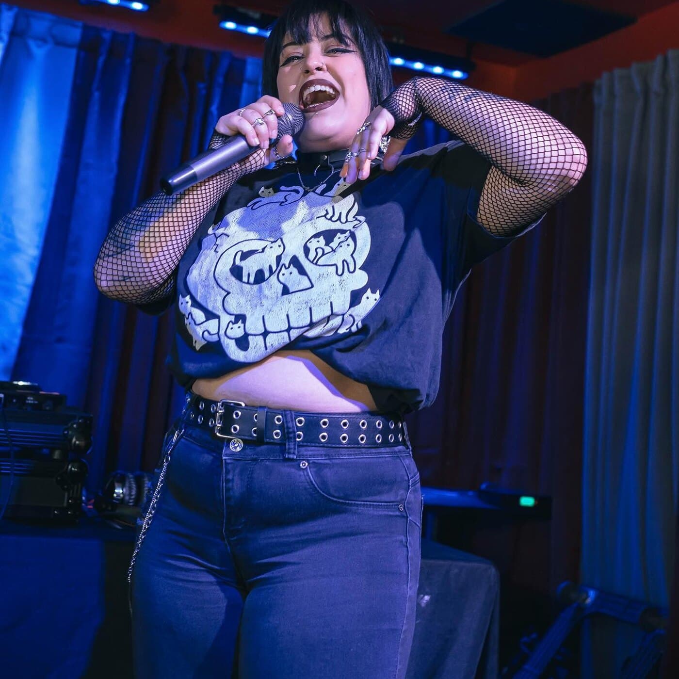 A singer in fishnet sleeves and a studded belt performs karaoke on stage under blue curtains at a Smells Like Sunday brunch