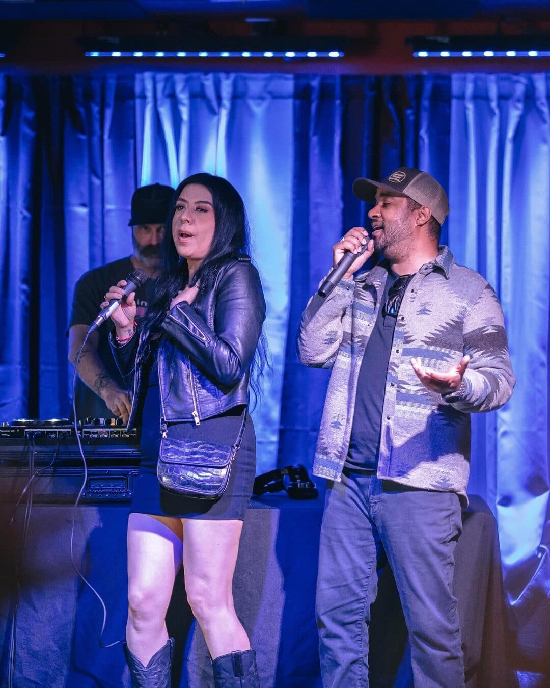 Two performers sharing the mic on stage in front of blue curtains and a DJ booth