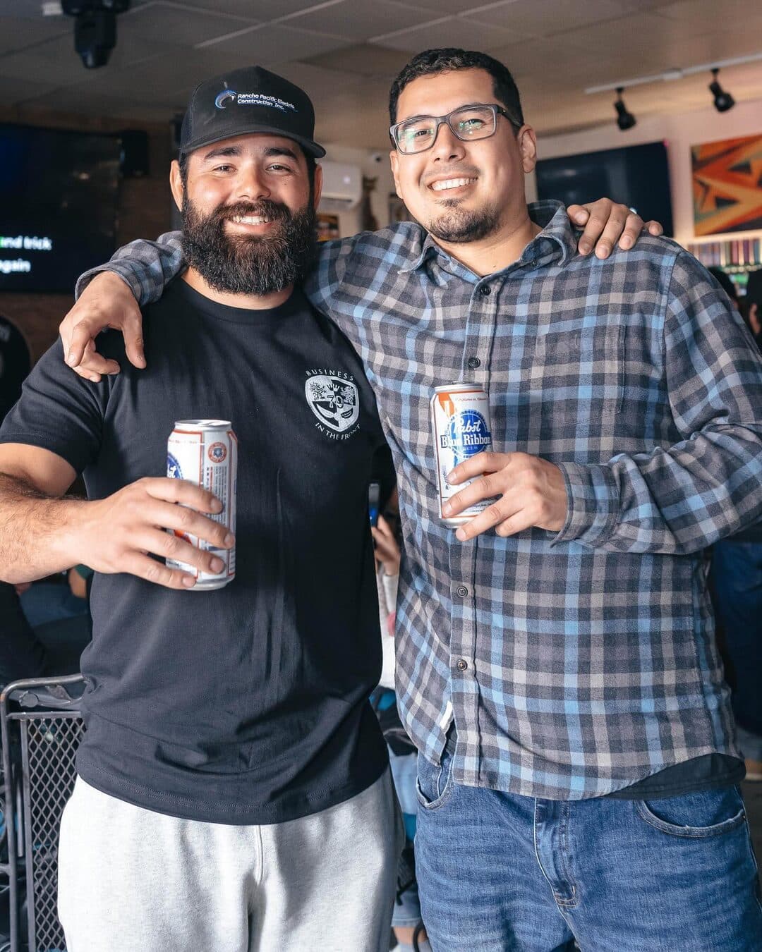 Two friends with their arms around each other, each holding a can of beer