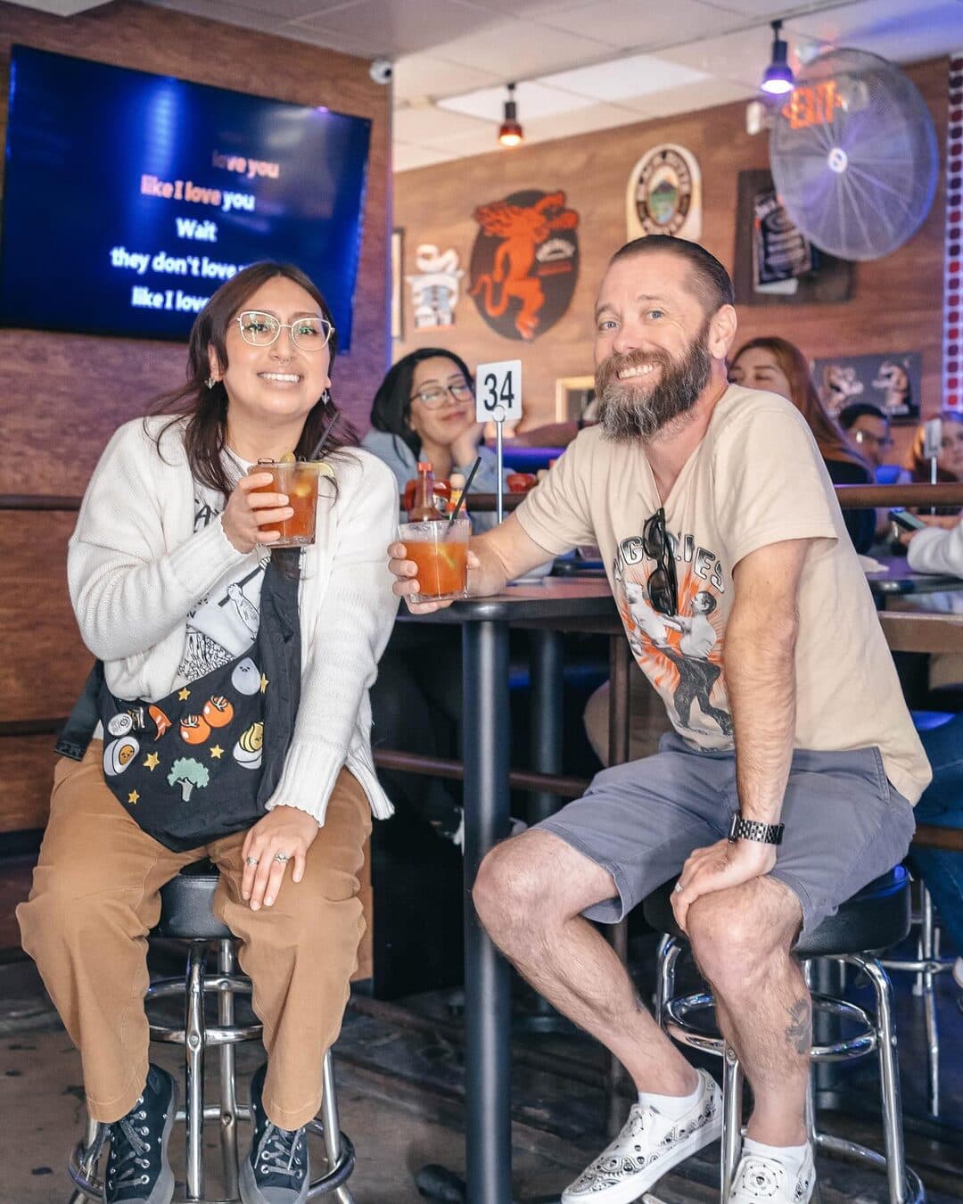 Two guests with drinks at a high-top table, a karaoke lyrics screen glowing behind them