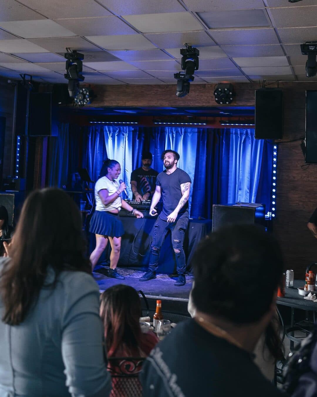 Wide view of the stage and room at a Smells Like Sunday brunch: a performer under blue curtains, guests at tables in the foreground